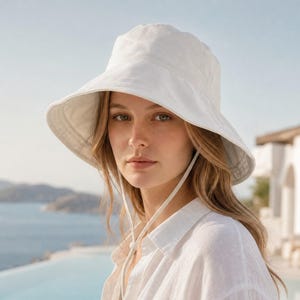 May include: A solid white bucket hat featuring a wide, downward-sloping brim and a matching white chin strap with an adjustable toggle. The hat is constructed from a smooth, lightweight fabric and provides full coverage for the head and face.