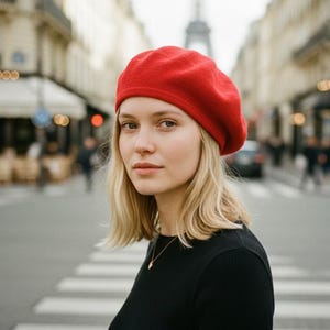 May include: A woman wearing a red beret and a black long-sleeve shirt stands on a crosswalk in front of the Eiffel Tower. The beret is a solid, vibrant red. The background shows Parisian architecture and street scenes.