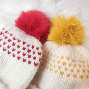 Peut inclure: Deux bonnets d'hiver tricotés avec des pompons duveteux. L'un a un pompon rouge et des motifs en forme de cœur rouges, l'autre a un pompon jaune et des motifs en forme de cœur jaunes. Les bonnets sont blancs et faits d'un fil épais et texturé.