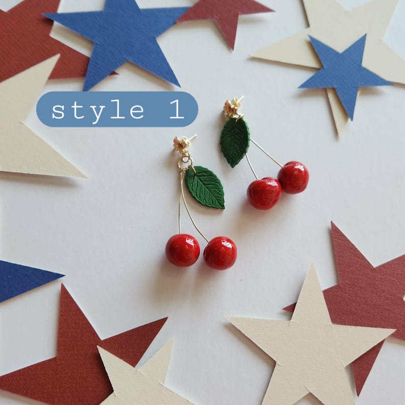 May include: A pair of dangle earrings featuring two glossy red cherry beads attached to a gold-tone metal stem with a textured green leaf. The text "style 1" is visible in a blue rounded rectangle above the earrings, surrounded by paper stars.
