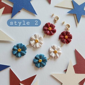 May include: A pair of dangle earrings featuring three stacked flower charms in red, white, and blue with yellow centers, connected by gold-toned metal links. The text "style 2" is visible in a blue rounded rectangle in the upper left corner.
