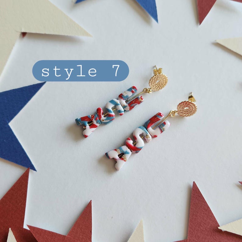 May include: A pair of dangle earrings featuring a marbled red, white, and blue pattern with gold-toned textured circular studs. The earrings are labeled style 7 in a blue rectangular box. The design consists of curved, multi-colored segments.