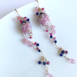 May include: A pair of delicate, floral-themed earrings. The earrings feature pink, purple, and blue beads, with a gold-colored wire. The design includes small pink flowers and teardrop-shaped beads. The earrings are on a white surface.