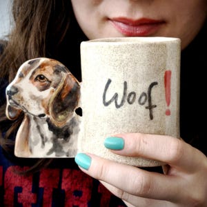以下が含まれることがあります： ベージュ色のセラミックマグカップ、ビーグル犬のハンドル付き。マグカップには「Woof！」の文字が黒と赤で書かれています。犬のハンドルは茶色、黒、白のディテールでペイントされています。