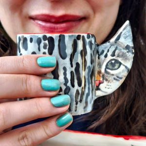 Puede incluir: Una taza de cerámica con un asa con forma de cara de gato y un estampado de manchas negras y grises. El interior de la taza es rojo. La taza la sostiene una persona con esmalte de uñas turquesa.