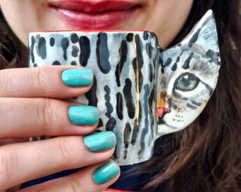 Puede incluir: Una taza de cerámica con un asa con forma de cara de gato y un estampado de manchas negras y grises. El interior de la taza es rojo. La taza la sostiene una persona con esmalte de uñas turquesa.