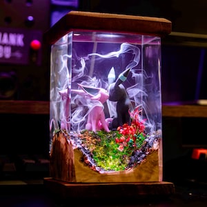 May include: A glass display case with a wooden base, featuring two small pink and black figurines of Pokemon characters, surrounded by a white smoke-like substance and green moss. The case is illuminated from within, creating a glowing effect.