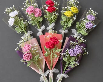 May include: A collection of handmade crochet flower bouquets in various colors, including white, pink, red, yellow, and purple. Each bouquet is wrapped in colorful paper and clear cellophane, tied with a white ribbon, and features delicate baby's breath.