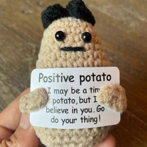 May include: A crocheted potato toy wearing a black hat holds a white card with the text "Positive potato I may be a tiny potato, but I believe in you. Go do your thing!"