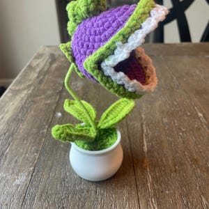 May include: A crocheted purple and green plant with a large open mouth, resembling a carnivorous plant. The plant is in a white pot and is sitting on a wooden table.