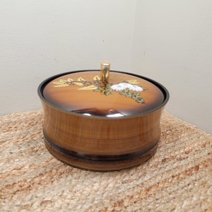 May include: A round wooden box with a gold knob on the lid. The lid is decorated with a floral design in shades of brown, white, and gold.