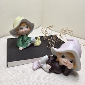 May include: Two ceramic elf figurines. One elf wears a green shirt and yellow hat and shoes. The other elf wears a brown shirt and a lavender hat and shoes. Both have freckles and are lying down.