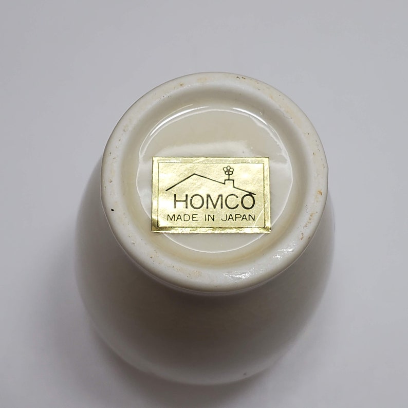 K&ouml;nnte beinhalten: Wei&szlig;e Keramiktasse mit einem goldenen Etikett mit der Aufschrift "HOMCO MADE IN JAPAN".