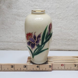 K&ouml;nnte beinhalten: Eine kleine, cremefarbene Keramikvase mit goldfarbenem Rand und floralem Design. Die Vase zeigt eine violette und rote Blume mit gr&uuml;nen Bl&auml;ttern.