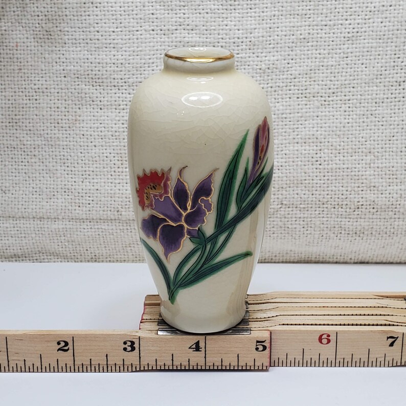 K&ouml;nnte beinhalten: Eine kleine, cremefarbene Keramikvase mit goldfarbenem Rand und floralem Design. Die Vase zeigt eine violette und rote Blume mit gr&uuml;nen Bl&auml;ttern.