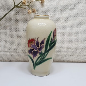 K&ouml;nnte beinhalten: Eine kleine, cremefarbene Keramikvase mit goldfarbenem Rand und einem Blumendesign. Die Vase zeigt eine violette und rote Blume mit gr&uuml;nen Bl&auml;ttern.