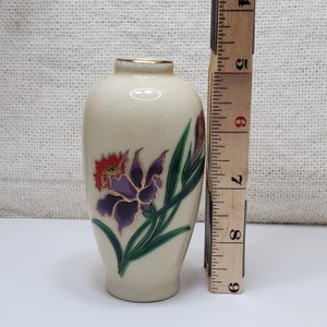 K&ouml;nnte beinhalten: Eine kleine, cremefarbene Keramikvase mit goldfarbenem Rand und einem Blumendesign. Die Vase zeigt eine violette und orangefarbene Blume mit gr&uuml;nen Bl&auml;ttern.