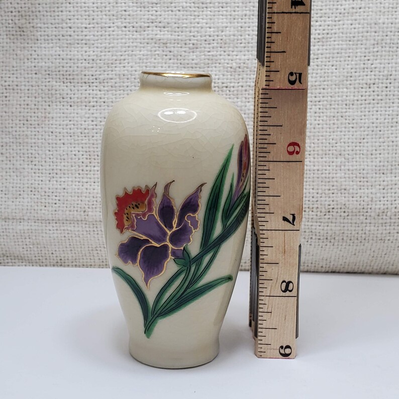 K&ouml;nnte beinhalten: Eine kleine, cremefarbene Keramikvase mit goldfarbenem Rand und einem Blumendesign. Die Vase zeigt eine violette und orangefarbene Blume mit gr&uuml;nen Bl&auml;ttern.