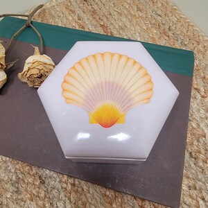 May include: A lavender hexagonal trinket box with a painted seashell design. The seashell is yellow, orange, and pink.