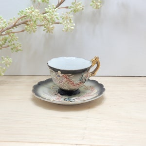 May include: A small, white teacup and saucer with a black and gold dragon design. The cup has a gold handle and the saucer has a scalloped edge.