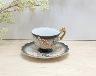 May include: A small, white teacup and saucer with a black and gold dragon design. The cup has a gold handle and the saucer has a scalloped edge.