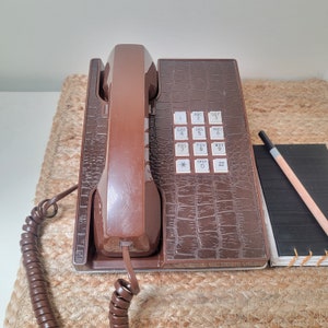May include: A vintage brown telephone with a textured, faux-leather finish. The phone has a keypad with white buttons and a coiled cord. A small notepad and pencil are next to the phone.