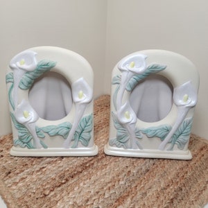 May include: Two white ceramic picture frames with a raised design of white calla lilies and green leaves. The frames are empty and ready for photos.