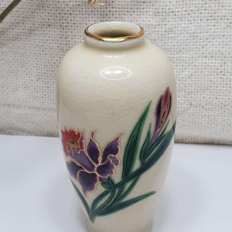 K&ouml;nnte beinhalten: Eine kleine, cremefarbene Keramikvase mit goldfarbenem Rand. Die Vase ist mit einem Blumendesign verziert, das lila, rote und gr&uuml;ne Blumen zeigt.