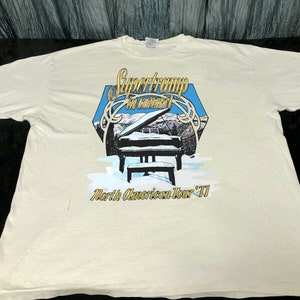 Puede incluir: Camiseta blanca con un gráfico vintage de Supertramp "In Concert" North American Tour '77. El gráfico presenta un piano en un paisaje nevado.