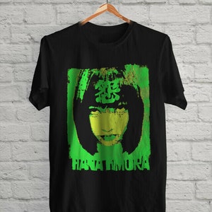 May include: Black t-shirt with a green and yellow graphic print of a woman's face with the text "HANAI MURA" underneath.