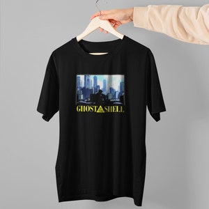 Könnte beinhalten: Schwarzes T-Shirt mit einem Grafikdruck einer Stadtlandschaft und dem Text "Ghost in the Shell" in Gelb.