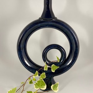 May include: A dark blue ceramic vase with a unique, abstract design. The vase features a large, circular opening and a smaller, twisted loop. The vase is perfect for displaying flowers or other decorative items.