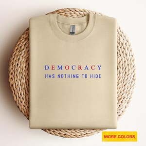Peut inclure: Un sweat-shirt beige avec l'inscription "DEMOCRACY HAS NOTHING TO HIDE" en bleu et rouge. Le sweat-shirt est posé sur un tapis rond et tissé. Les mots "MORE COLORS" sont dans un rectangle jaune en bas à droite.