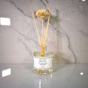 May include: A clear glass reed diffuser bottle containing liquid, featuring a white label with black text reading Retreat, and topped with several thin wooden reeds and a single cream-colored decorative flower at the top of the central reed.