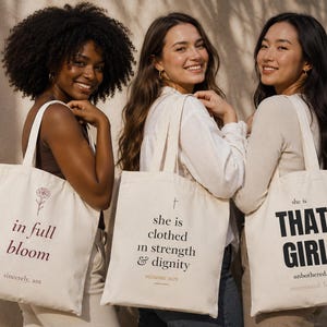 May include: Three women smiling, each holding a cream-colored tote bag. The bags feature text and floral designs in dark red and black. One bag reads "in full bloom," another "she is clothed in strength & dignity," and the third says "THAT GIRL."