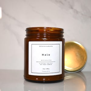May include: An amber glass jar candle with a white label featuring the text #SincerelyAntCo, Halo, White Eucalyptus, All Natural / Non-Toxic, Soy Wax Candle, and 8 oz / 230 g. A gold-colored metal lid rests to the right of the candle jar.