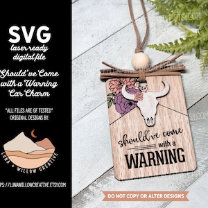 May include: A wooden car charm featuring a white skull and floral design. The charm hangs from a brown cord and has the text "Should've come with a WARNING" printed on it.