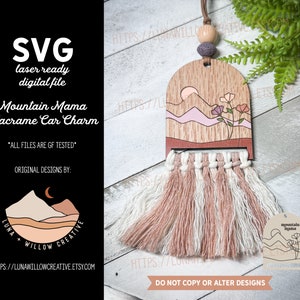 May include: A wooden mountain mama macrame car charm with a pink and white macrame tassel. The wooden charm features a sunset over mountains with pink and orange flowers. The text "mountain mama" is on a separate tag.