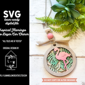 Könnte beinhalten: Ein zweilagiger Holz-Autoanhänger mit einem rosa Flamingo und grünen tropischen Blättern. Der Anhänger ist rund und hat oben ein Loch zum Aufhängen. Der Anhänger ist aus lasergeschnittenem Holz und kann bemalt oder gebeizt werden.