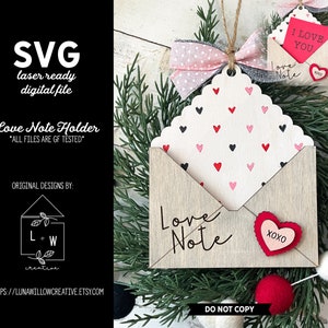 May include: A wooden love note holder shaped like an envelope with a white background and a pattern of red and black hearts. The envelope has a flap that opens to reveal a space for a note. The words "Love Note" are written on the front of the envelope. The holder is decorated with a red heart with the word "XOXO" on it.