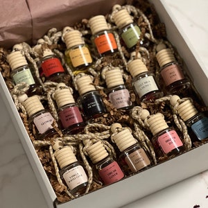 May include: A white box filled with small glass bottles with cork stoppers and string. The bottles are filled with colorful liquids and have labels with various scents, such as "Vanilla White Cove", "Dream Lover", "Glow Up", "Sunday Morning", and "Milk & Cereal".