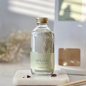May include: A clear glass bottle with a gold lid filled with a reed diffuser. The bottle has a white label with the text "SPA DAY" and the scent description "white tea, ginger and citrus". The bottle is sitting on a white marble surface with a few red rose petals scattered around it. The text "reed diffuser 100 ml" is also on the label.