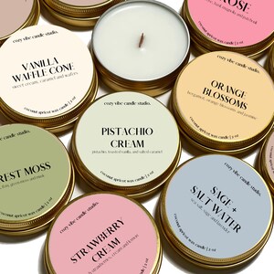 May include: A collection of small, round candles in gold tins. Each candle has a label with a different scent name, such as "Vanilla Waffle Cone" and "Pistachio Cream." The labels also include the candle's weight, 2 oz.