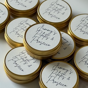 May include: A collection of round gold metal tins with white circular labels on the lids. Each label features the black handwritten text "Jummy's Pilates & Prosecco" in a cursive script. The tins are arranged in a cluster, showing the metallic finish.