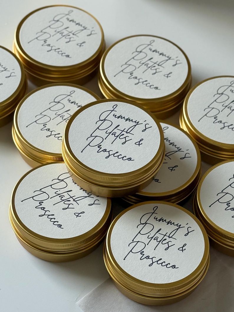 May include: A collection of round gold metal tins with white circular labels on the lids. Each label features the black handwritten text "Jummy's Pilates & Prosecco" in a cursive script. The tins are arranged in a cluster, showing the metallic finish.