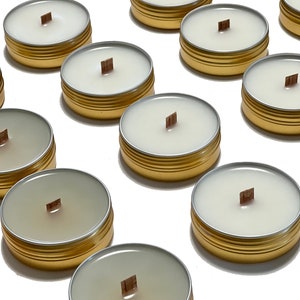 May include: Several small, round candles in gold-colored tins with wooden wicks. The candles have a creamy white wax and are arranged on a white surface. The lids are visible in the background. The overall theme is home fragrance.