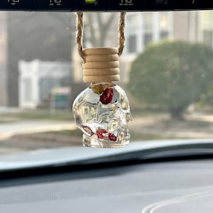 May include: A clear glass skull-shaped car air freshener with a wooden top and red and yellow dried flowers inside. The air freshener is hanging from a rearview mirror.