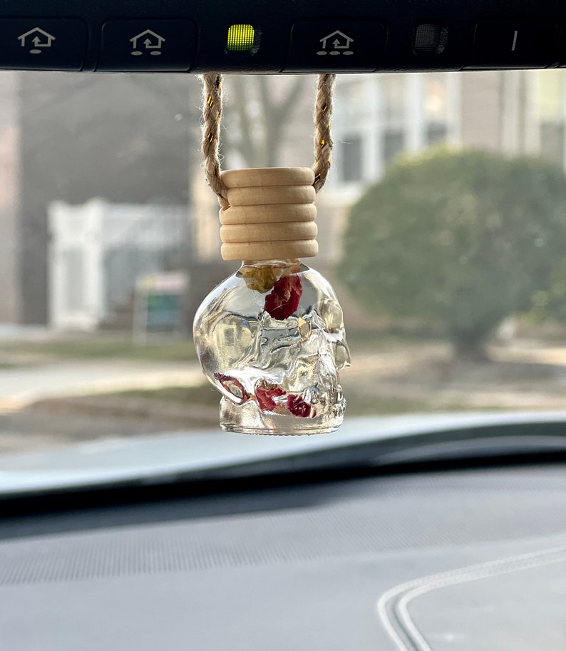 May include: A clear glass skull-shaped car air freshener with a wooden top and red and yellow dried flowers inside. The air freshener is hanging from the rear-view mirror of a car.