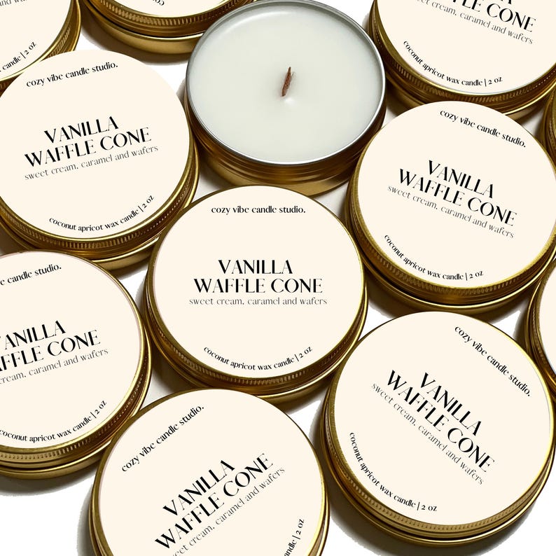 May include: A collection of small, round, gold-colored tin candles with white wax and wooden wicks. The labels read "VANILLA WAFFLE CONE" with "sweet cream, caramel and wafers" below. The text "cozy vibe candle studio" and "coconut apricot wax candle 2 oz" are also visible.