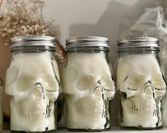 Könnte beinhalten: Drei durchsichtige Glasgefäße in Form von menschlichen Schädeln, gefüllt mit weißem Wachs. Die Gefäße haben silberfarbene Schraubdeckel aus Metall. Die schädelförmigen Glasbehälter zeigen detaillierte Augenhöhlen, Nasenlöcher und Zähne.
